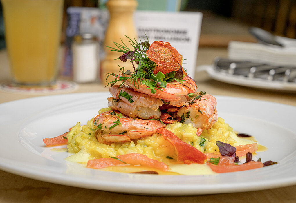 Risotto im Conviva Blaues Haus Ein Teller mit cremigem Risotto, darauf mehrere gebratene Garnelen, garniert mit frischen Kräutern und roten Chips.